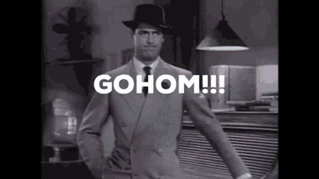 a man in a suit and hat is standing in a room with the words gohom !!! written on the screen .
