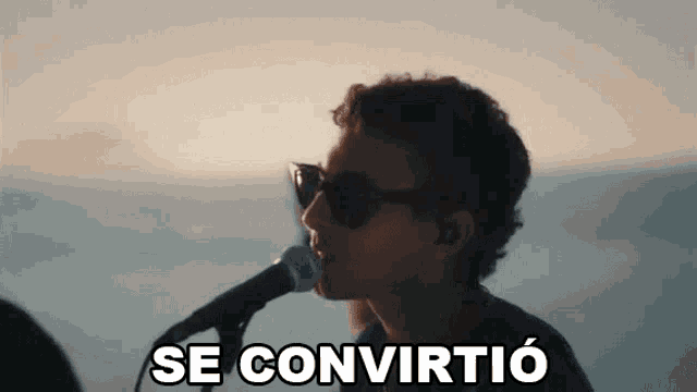 a man wearing sunglasses is singing into a microphone with the words `` se convirtio '' written below him .