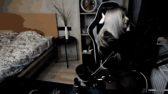 a woman is sitting in a gaming chair in a bedroom with a bed in the background .