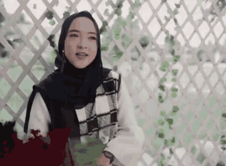 a woman wearing a hijab and a plaid vest is standing in front of a white lattice fence .