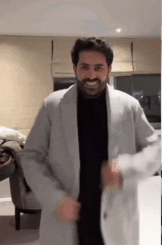 a man with a beard is standing in a living room wearing a coat and smiling .