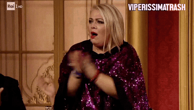 a woman wearing a purple sequined cape applauds in front of a sign that says viperissima trash