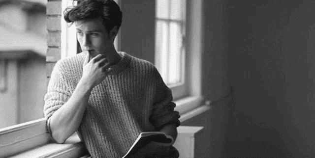a young man is leaning against a window sill while reading a book .