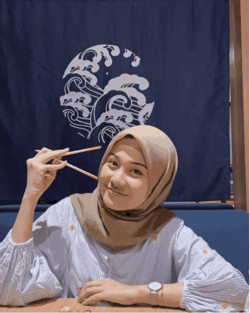 a woman wearing a hijab holds chopsticks over her mouth
