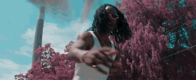 a man wearing sunglasses and a white tank top is dancing in front of a purple tree .