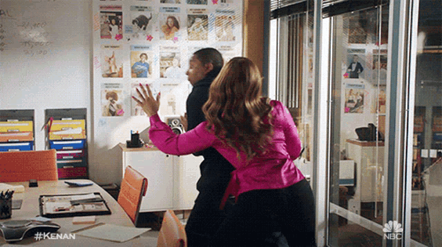 a woman in a pink shirt is standing next to a man in an office with a nbc logo on the wall