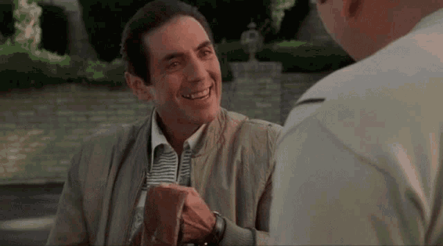 a man in a jacket is smiling while holding a cup of coffee and talking to another man .