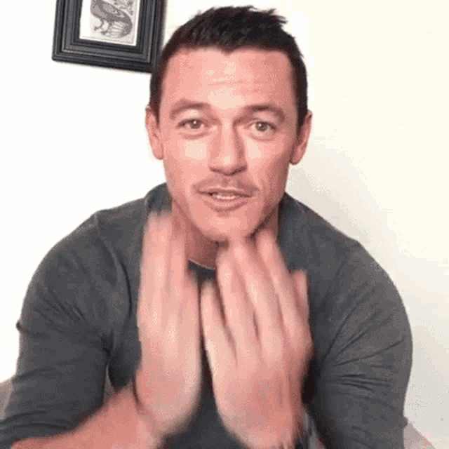 a man in a gray shirt is clapping his hands together in front of his face .