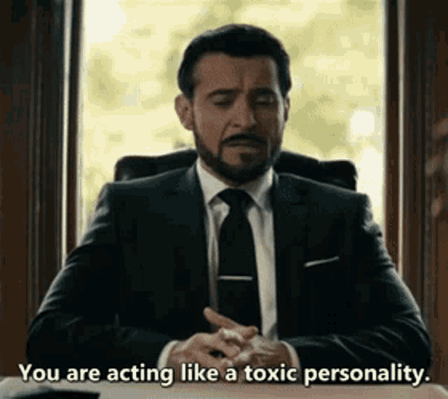 a man in a suit and tie is sitting in front of a window and saying you are acting like a toxic personality .