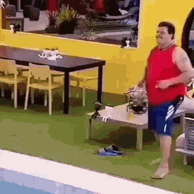 a man in a red tank top and blue shorts is standing in front of a table and chairs .