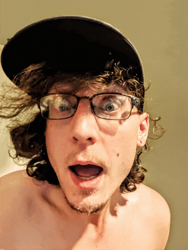 a shirtless man wearing glasses and a hat looks surprised