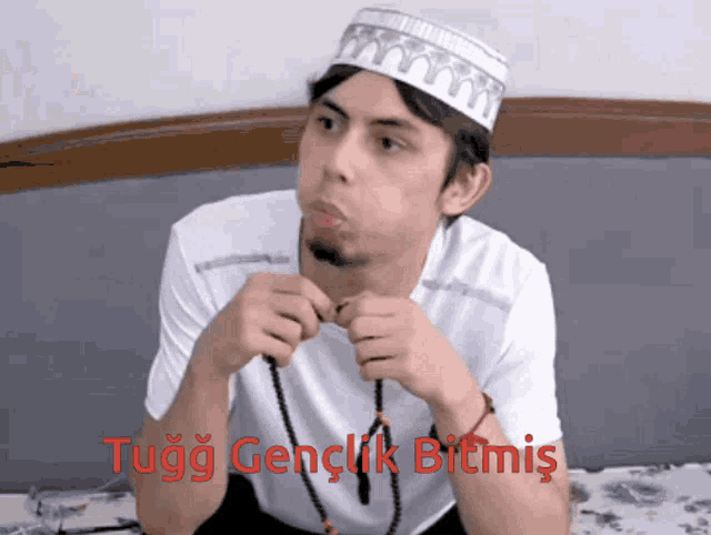 a man wearing a white hat is sitting on a bed with the words tugg genclik bitmis written above him