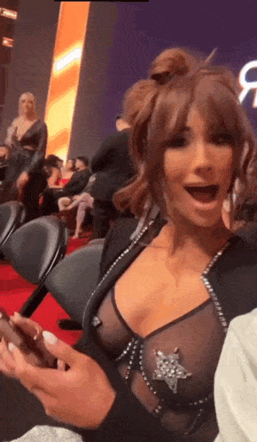 a woman in a sheer bra is sitting on a red carpet and looking at her phone