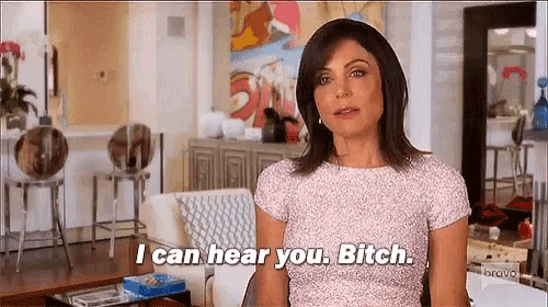 a woman in a pink shirt is standing in a living room and says i can hear you bitch .