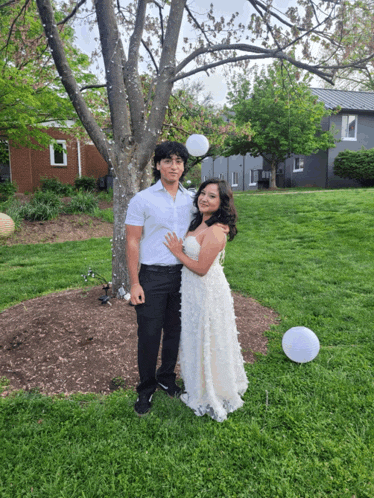 a man in a white shirt and a woman in a white dress pose for a picture in front of a tree