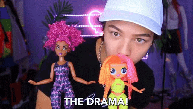 a man in a white hat holds two dolls and says " the drama "