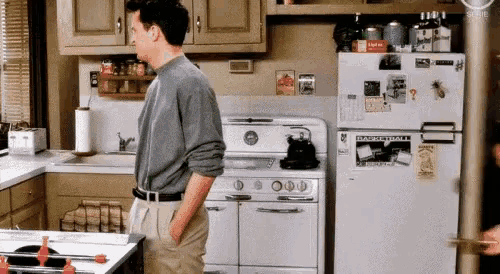 a man is standing in a kitchen with his hands in his pockets and a refrigerator with a sticker on it .