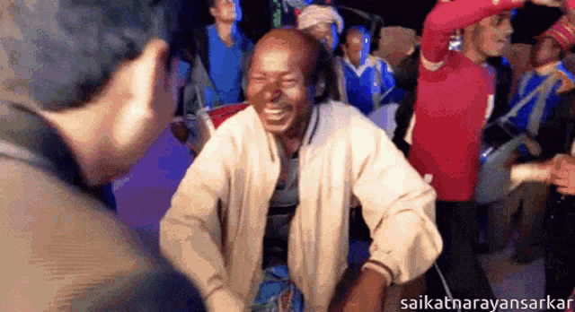 a man in a white jacket is laughing in front of a group of people with the name saikatarayansarkar at the bottom