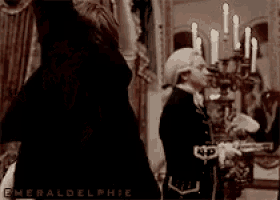 a man in a wig is standing in a room with candles and a chandelier .