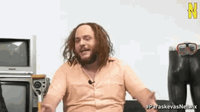 a man with long hair and a beard is sitting in front of a television and a sign that says paraskevasnetwork
