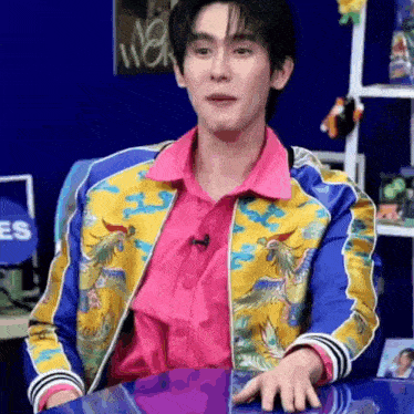 a young man in a colorful jacket and pink shirt is sitting at a table .