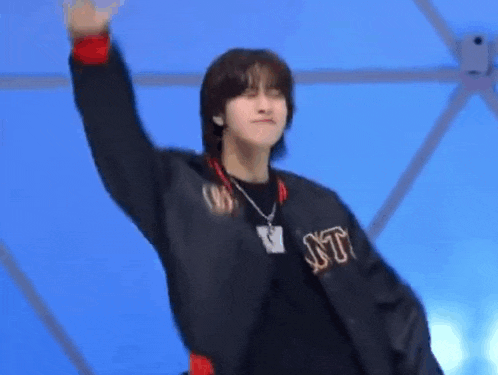 a young man wearing a black jacket and a necklace is waving his hand in the air .