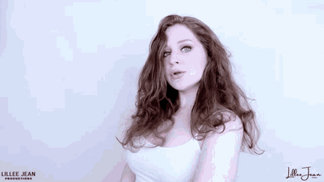 a woman with red hair and blue eyes is wearing a white tank top and looking at the camera .