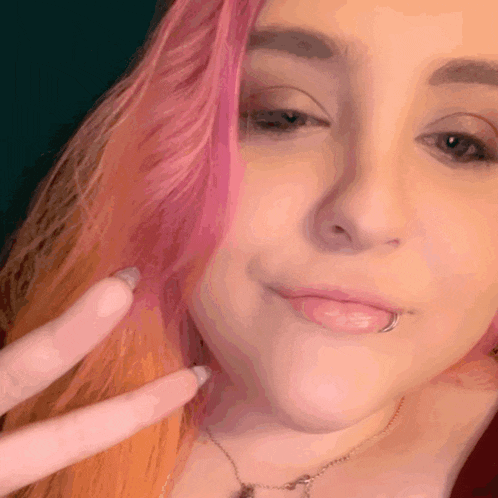 a close up of a woman 's face with pink hair making a peace sign
