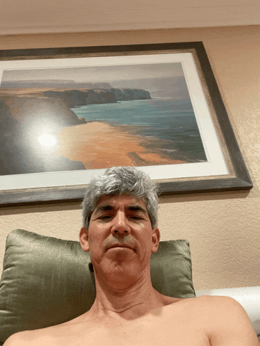 a man without a shirt is laying on a bed in front of a painting of the ocean