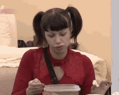 a woman in a red shirt is sitting on a couch eating food from a plastic container .