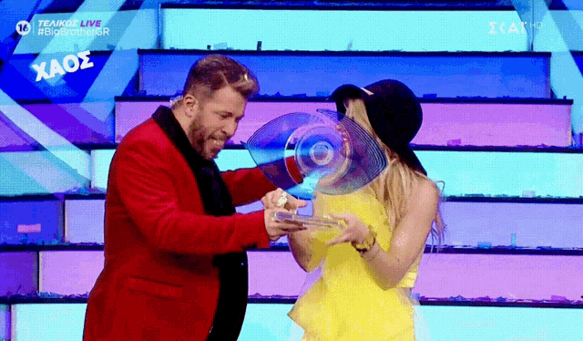 a man in a red suit is holding a trophy next to a woman in a yellow dress and a sign that says xaos
