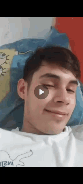 a young man is smiling while laying in bed with a play button on his face .