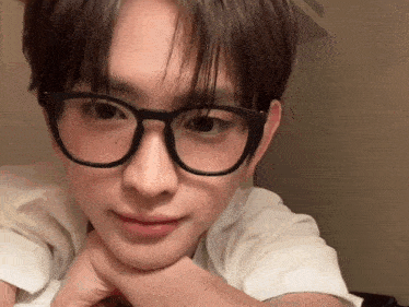 a young man wearing glasses and a white shirt is laying down with his hands on his chin .