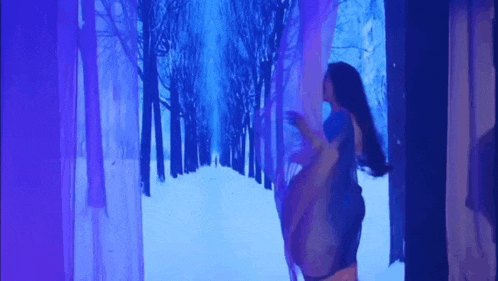 a woman in a blue dress is standing in front of a blue screen with a snowy forest projected on it .