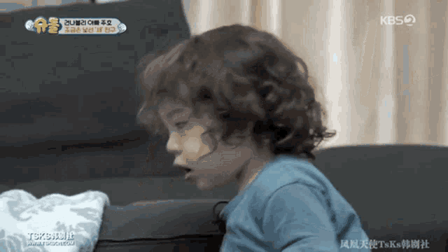 a little boy with curly hair is sitting on a couch in front of a screen that says kbs