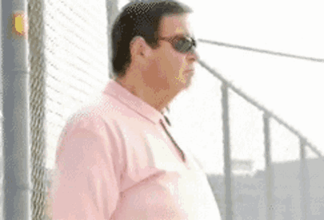 a man wearing sunglasses and a pink shirt is standing next to a chain link fence .