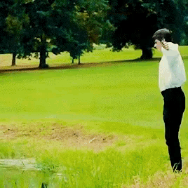 a man in a white shirt and black pants stands in the grass near a body of water