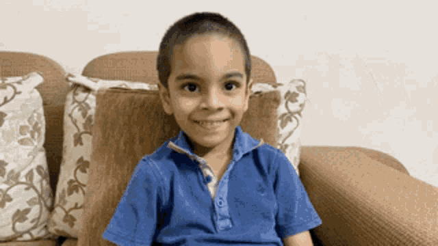 a young boy in a blue shirt sits on a couch smiling