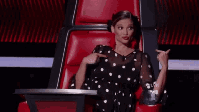 a woman in a black and white polka dot dress is sitting in a red chair and pointing at herself .