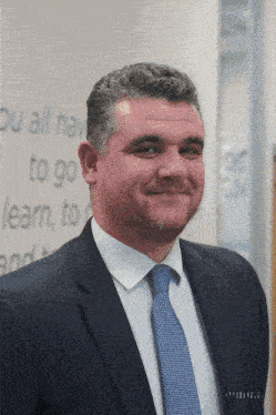 a man in a suit and tie smiles in front of a wall that says " you all have to go learn to "