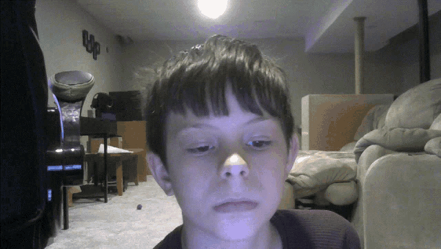 a young boy looks at the camera in a living room with a sign on the wall that says u.p.
