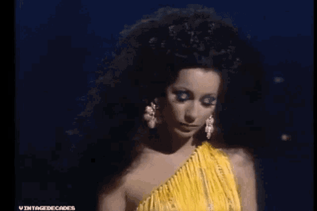 a woman with long black hair is wearing a yellow one shoulder dress and earrings .