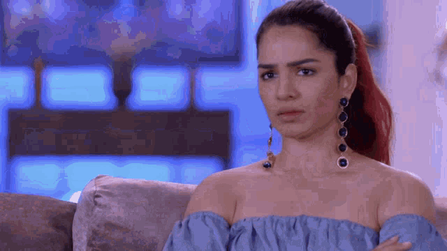 a woman in a blue off the shoulder top and earrings is sitting on a couch with her arms crossed .