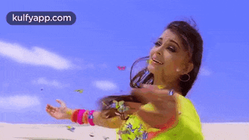 a woman in a yellow saree is standing on a beach with her arms outstretched and butterflies flying around her .