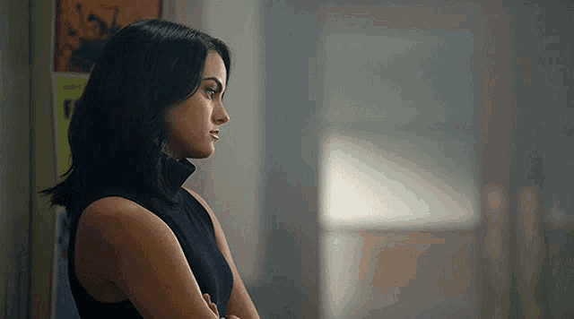 a woman in a black dress is standing with her arms crossed and looking out a window .