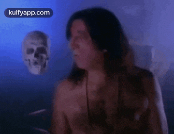 a man without a shirt is standing in front of a skull in a dark room .