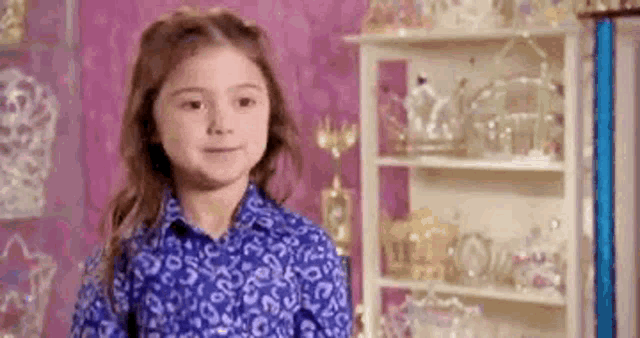 a little girl in a blue shirt is standing in front of a shelf full of trophies .