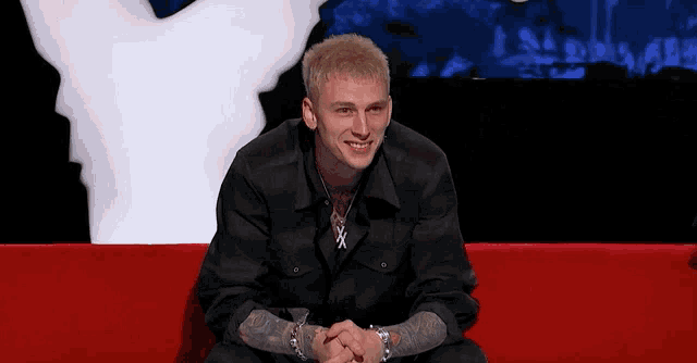 a man in a black shirt is sitting on a red couch with his hands folded and smiling .