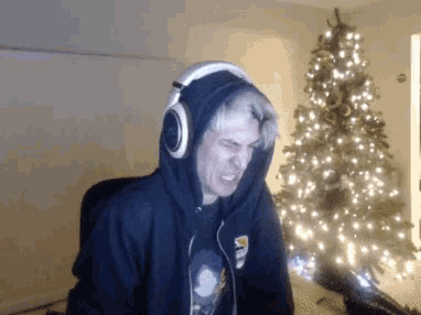 a man wearing a hoodie and headphones stands in front of a lit christmas tree