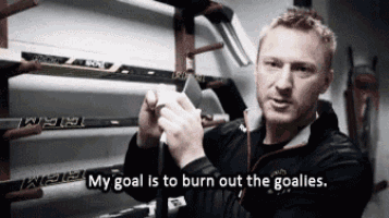 a man is holding a hockey stick in front of a rack of hockey sticks with the words my goal is to burn out the goalies